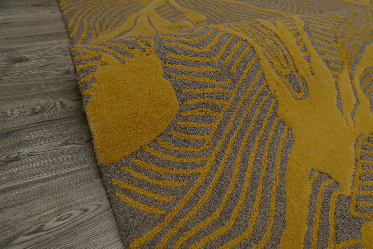 A close-up of a textured area rug with a mustard yellow and gray abstract leaf or wave pattern, shown laid out on a gray wood-plank floor.