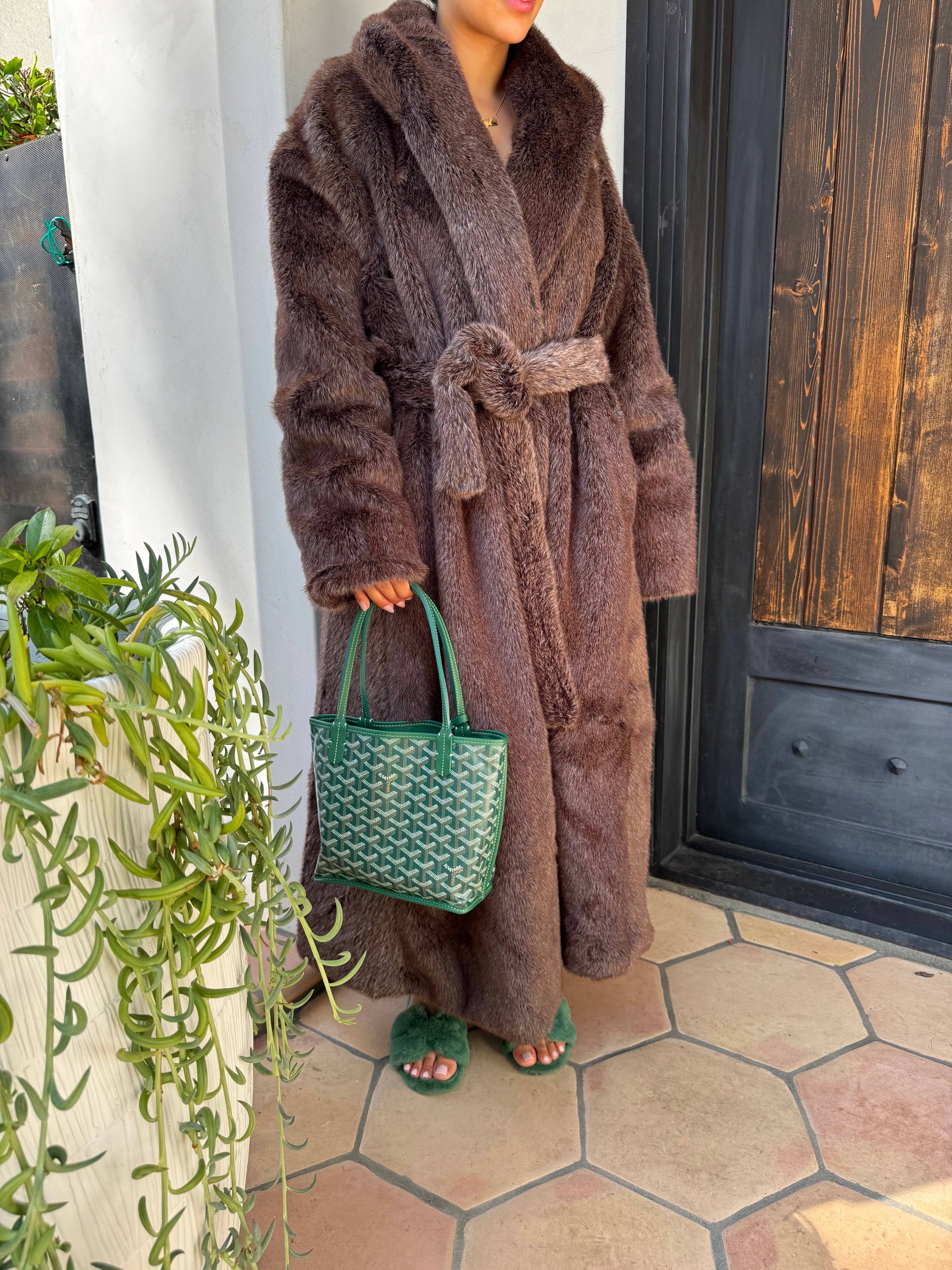 Brown plush robe with green textured bag on a white surface near a wooden door and tiled wall.