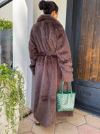 Person wearing a brown robe holding a green bag, standing in front of a door with plants on the floor.